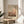 May include: Framed art print with abstract warm-toned composition in orange, pink and brown hues displayed on round wooden side table next to beige upholstered chair. Soft natural lighting creates gentle shadows across neutral wall. Green plant leaves visible in corner. Print features Mark Rothko's name at bottom in clean typography within thin gold or brass frame with cream matting.