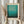 May include: Person in cream sweater holding framed poster featuring turquoise and teal gradient abstract art with museum attribution text below. Light wood frame with cream matting. Dried pampas grass stems held in other hand against soft neutral background with natural window lighting.