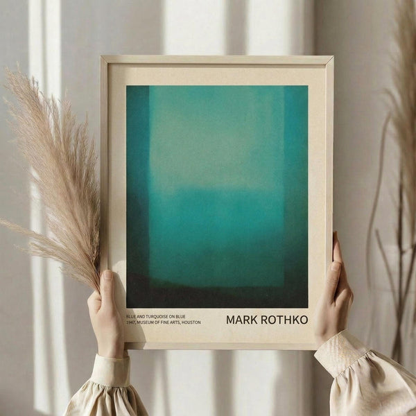May include: Person in cream sweater holding framed poster featuring turquoise and teal gradient abstract art with museum attribution text below. Light wood frame with cream matting. Dried pampas grass stems held in other hand against soft neutral background with natural window lighting.