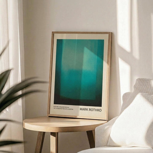May include: Framed art print displaying an abstract composition of turquoise and teal blue gradients with darker edges, sitting on a light wood round side table. The thin wood frame has a cream mat border with text identifying the artwork and artist. Scene shows part of a beige upholstered chair, plant leaves casting shadows on white walls, and natural sunlight streaming across the minimalist interior.