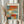 May include: Person in beige ruffled sleeves holding framed abstract art print featuring soft rectangular color blocks in orange, red, blue, teal, yellow and cream. Light wood frame against white curtained window with dried pampas grass stems visible to the left.