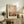 May include: Abstract watercolor artwork in warm earth tones displayed in light wooden frame on rustic wooden sideboard, surrounded by potted fern, ceramic vase, stacked books, and brown leather armchair in cozy living room setting with cream walls and soft natural lighting.