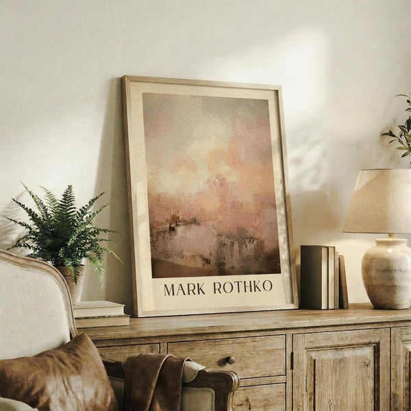 May include: Abstract watercolor artwork in warm earth tones displayed in light wooden frame on rustic wooden sideboard, surrounded by potted fern, ceramic vase, stacked books, and brown leather armchair in cozy living room setting with cream walls and soft natural lighting.