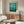 May include: Framed teal and turquoise gradient abstract art print displayed above wooden sideboard in minimalist living room with white curved chair, glass coffee table, and modern brass floor lamp. Print features soft blue-green color field with subtle geometric forms and museum exhibition text at bottom.