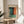 May include: Framed abstract art print in natural wood frame displaying soft color gradients of teal, orange, and green tones, displayed on round wooden side table next to beige upholstered chair with white throw pillow, green plant leaves visible in corner, warm natural lighting creating shadows on neutral wall