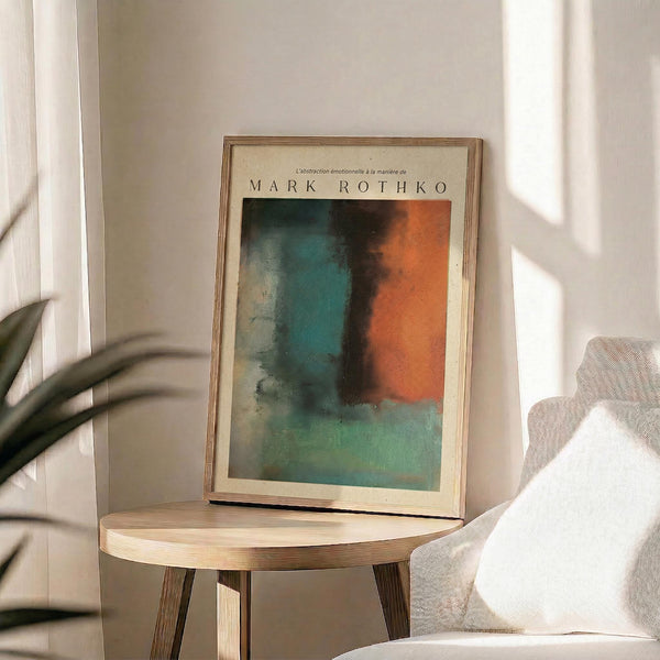 May include: Framed abstract art print in natural wood frame displaying soft color gradients of teal, orange, and green tones, displayed on round wooden side table next to beige upholstered chair with white throw pillow, green plant leaves visible in corner, warm natural lighting creating shadows on neutral wall