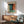 May include: Framed abstract art print with orange, teal, and green color blocks in the style of Mark Rothko, displayed above a mid-century modern wooden credenza with white table lamp, next to a gray upholstered armchair with wooden arms in a sunlit room with white walls