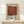 May include: Framed poster featuring Mark Rothko's name in bold black text above a large rust-red square, with exhibition details and descriptive text below on cream background. Natural wood frame sits on light wooden surface beside small white wrapped gift with evergreen sprig against neutral wall.