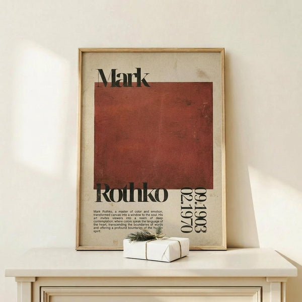 May include: Framed poster featuring Mark Rothko's name in bold black text above a large rust-red square, with exhibition details and descriptive text below on cream background. Natural wood frame sits on light wooden surface beside small white wrapped gift with evergreen sprig against neutral wall.