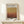 May include: Framed Mark Rothko exhibition poster with earth-toned gradient artwork in warm browns and golden hues, displayed on light wood shelf against cream wall. Small wrapped gift with twine and evergreen sprig sits beside the wooden-framed print. Soft natural lighting creates gentle shadows on the minimalist wall display.