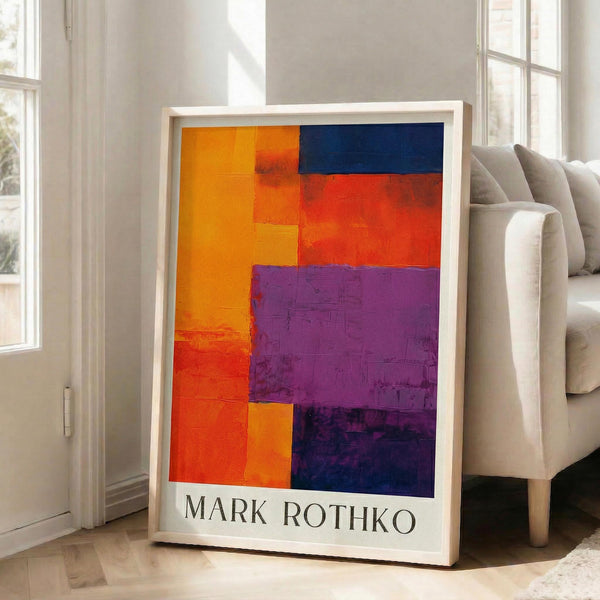 May include: Framed abstract art print with bold geometric color blocks in orange, red, purple, and blue arranged in rectangular sections. The poster features "MARK ROTHKO" text at bottom in black letters. Shown leaning against white wall next to beige upholstered chair in bright living room with large windows and hardwood floors.
