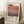 May include: Framed art print leaning against white wall in cozy room setting. Poster features abstract gradient painting in warm coral, pink and purple horizontal bands with "Mark Rothko" text overlay and exhibition details. Light wood frame, positioned next to wooden stool with white coffee cup, textured cream rug, and soft natural lighting creating window shadows on walls.