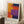 May include: Abstract colorful artwork print in light wood frame leaning against white wall corner. Features bold geometric blocks in orange, red, purple, and navy blue with visible paint texture. Room setting shows white tea cup on wooden block, cream throw blanket, and fluffy white rug. Natural window light creates soft shadows on wall.