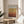 May include: Framed poster with Mark Rothko exhibition text and gradient artwork in golden yellow fading to deep reddish-brown horizontal color blocks, displayed on light wood round side table next to beige upholstered chair with white pillow, green plant leaves visible, soft natural lighting creating shadows on cream wall background.