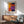 a modern living room with a wooden cabinet, a green armchair, and a wooden coffee table. Above the cabinet, there is a framed abstract painting with a vibrant mix of orange, purple, and yellow colors.