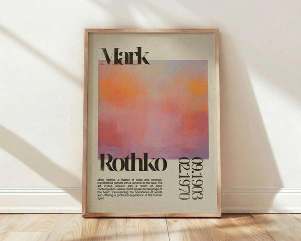 May include: Framed art print featuring Mark Rothko's name in bold black text above a gradient abstract painting with orange, pink, and purple hues. Below the artwork are dates and descriptive text about the artist. The print has a cream-colored background and sits in a light wood frame, leaning against a white wall on light wooden flooring with soft natural lighting casting shadows.