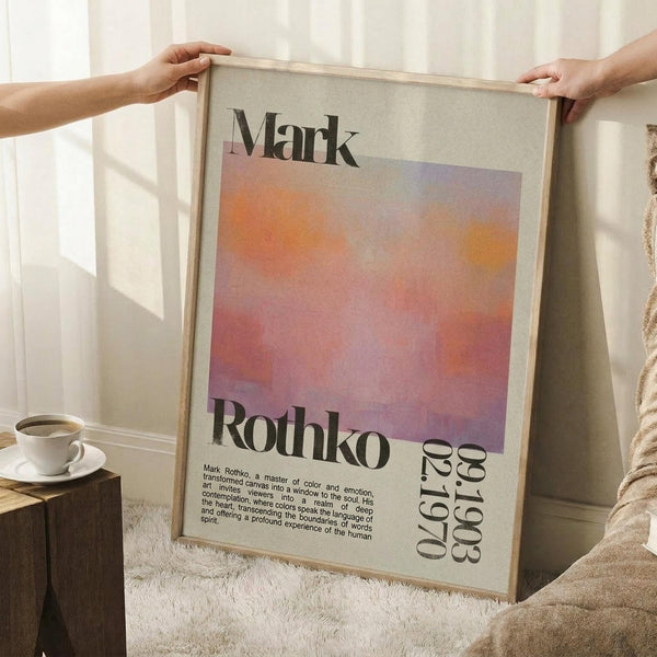 May include: Person holding large framed poster featuring abstract orange and purple gradient artwork with "Mark Rothko" text and biographical description. Light wooden frame, white matting, displayed in bright room with coffee cup on wooden surface and soft textiles nearby.