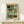 May include: Abstract art print in thin wooden frame leaning against white wall on light wood surface. Artwork features muted blue-green and golden yellow geometric blocks with black accents in painterly style. Small white gift box with natural twine and dried sprig positioned in front of frame.