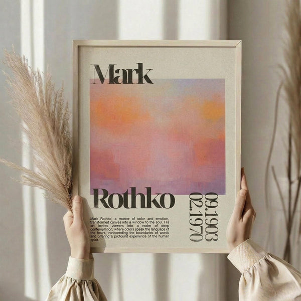 May include: Person in cream sweater holding framed poster featuring orange and purple gradient watercolor design with "Mark Rothko" text and biographical description. Light wooden frame against neutral curtained background with dried pampas grass visible to left side.