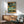May include: Abstract painting with geometric blocks in teal, sage green, orange, and black tones displayed above a mid-century modern wooden sideboard. Contemporary living room setting features a mint green upholstered armchair with wooden frame, small round coffee table, and ceramic table lamp. Warm natural lighting creates shadows on white wall.