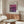 May include: Modern living room displaying framed Mark Rothko gallery exhibition poster with purple and pink abstract artwork. Poster hangs above mid-century wooden credenza with four cabinet doors. Scene features white curved chair, glass oval coffee table, brass floor lamp with circular mirror detail, and white table lamp. Neutral beige walls and natural lighting create minimalist Scandinavian aesthetic.