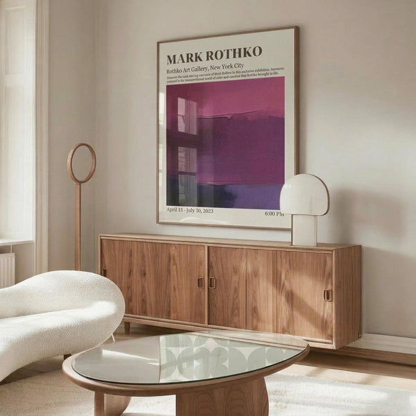 May include: Modern living room displaying framed Mark Rothko gallery exhibition poster with purple and pink abstract artwork. Poster hangs above mid-century wooden credenza with four cabinet doors. Scene features white curved chair, glass oval coffee table, brass floor lamp with circular mirror detail, and white table lamp. Neutral beige walls and natural lighting create minimalist Scandinavian aesthetic.