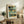 May include: Abstract art print with blue-green and golden yellow geometric blocks displayed in light wood frame on rustic wooden sideboard. Room setting shows fern plant, ceramic vase, books, and beige armchair with brown leather pillow. Soft natural lighting creates warm, modern living space atmosphere.