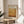May include: Framed poster featuring Mark Rothko exhibition details and warm golden-orange abstract artwork, displayed on round wooden side table next to beige upholstered chair with white pillow. Natural lighting creates soft shadows on neutral wall, with green plant leaves visible in corner of minimalist living room setting.