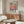 May include: Modern living room with cream walls featuring framed Mark Rothko exhibition poster with gradient orange, pink and purple color field. Wood credenza below holds minimalist table lamp. White curved chair and oval glass coffee table in foreground. Natural lighting creates warm, contemporary atmosphere showcasing mid-century modern furniture and art poster display.
