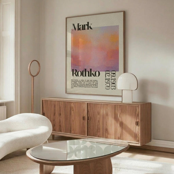 May include: Modern living room with cream walls featuring framed Mark Rothko exhibition poster with gradient orange, pink and purple color field. Wood credenza below holds minimalist table lamp. White curved chair and oval glass coffee table in foreground. Natural lighting creates warm, contemporary atmosphere showcasing mid-century modern furniture and art poster display.