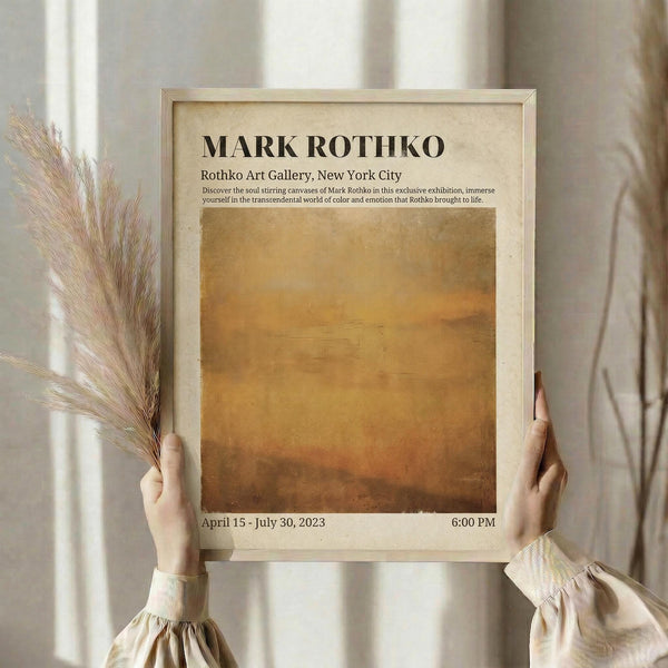 May include: Hands holding a framed exhibition poster featuring a warm golden-brown gradient artwork. The cream-colored frame displays Mark Rothko gallery information with exhibition dates April 15 - July 30, 2023. Person wears a beige blouse with gathered sleeves while holding dried pampas grass stems against a soft neutral curtained background.
