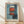 May include: Framed abstract painting poster in wooden frame with white matting, leaning against white wall on light wooden floor. Artwork shows blue-toned background with geometric red-orange rectangular shapes and white accents in painterly style. Natural window light creates soft shadows across the scene.
