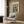 May include: Modern living room scene with cream armchair featuring geometric patterned pillow, brass floor lamp with white shade, dark round side table with open book, and wood-paneled wall displaying framed abstract art poster with colorful geometric squares in teal, orange, yellow and burgundy tones.