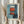 May include: Person holding framed abstract art print in light wood frame against white curtained window. Artwork shows orange-red rectangular forms with white elements on blue background in painterly style. Dried pampas grass held in other hand. Soft natural lighting on cream colored clothing.