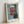 May include: Framed abstract art print in light wood frame leaning against white wall. Blue-teal background with geometric orange-red rectangular shapes stacked vertically, separated by white and dark horizontal bands. Soft, blurred brushstroke style with muted colors. Print includes artist attribution text at bottom. Frame casts subtle shadow on wall.