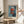 May include: Framed Mark Rothko abstract art print displayed on round wooden side table next to beige upholstered chair. Hand reaches toward the artwork which features stacked red-orange rectangular forms against blue background. Natural lighting creates soft shadows on neutral wall. Green plant leaves visible in corner of minimalist living room setting.