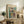 May include: Abstract art poster with geometric squares in teal, orange, yellow, and purple tones displayed in light wood frame on rustic wooden sideboard. Room styled with fern plant, cream ceramic vase, stacked books, and beige upholstered chair with brown throw pillow against white wall.