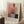May include: Framed abstract art poster with geometric blocks in warm coral pink, deep red, orange, gray, and dark green colors on cream background, leaning against white wall corner. Wooden frame, white mat border, artist name at bottom. Cozy room setting with coffee cup on wooden table, cream textured rug, and beige throw blanket visible.