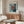 May include: Modern living room with wooden credenza displaying framed Bauhaus exhibition poster featuring Mark Rothko's abstract painting with red and orange rectangular forms on blue background. White sculptural chair, glass coffee table, and minimalist floor lamp complete the mid-century modern interior styling.