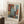 May include: Abstract art poster with geometric blocks in rust orange, dusty blue, cream, and sage green colors displayed in light wood frame, leaning against white wall in cozy room setting with coffee cup on wooden side table, textured cream rug, and beige throw blanket visible.
