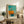 May include: Framed abstract art print displayed on rustic wooden sideboard featuring turquoise sky, orange geometric sailboat shape, and water reflection below. Print has light wood frame with white matting and "MARK ROTHKO" text at bottom. Scene includes fern plant, cream ceramic vases, books, and beige upholstered furniture in warm-toned living room setting.