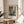 May include: Abstract art print in wooden frame displayed on round wooden side table next to beige upholstered chair. The artwork features geometric rectangular blocks in warm orange, rust red, teal blue, sage green, and cream colors with soft, painterly edges. Natural sunlight streams across the neutral-toned room setting with white curtains and plant leaves visible in corner.