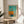 May include: Framed abstract art print displayed on round wooden side table next to beige upholstered chair. Artwork features turquoise and orange geometric shapes resembling a sailboat against blue-green sky with water reflection below. Natural sunlight streams across white wall creating soft shadows from nearby plant leaves. Light wood frame complements minimalist Scandinavian-style interior setting.