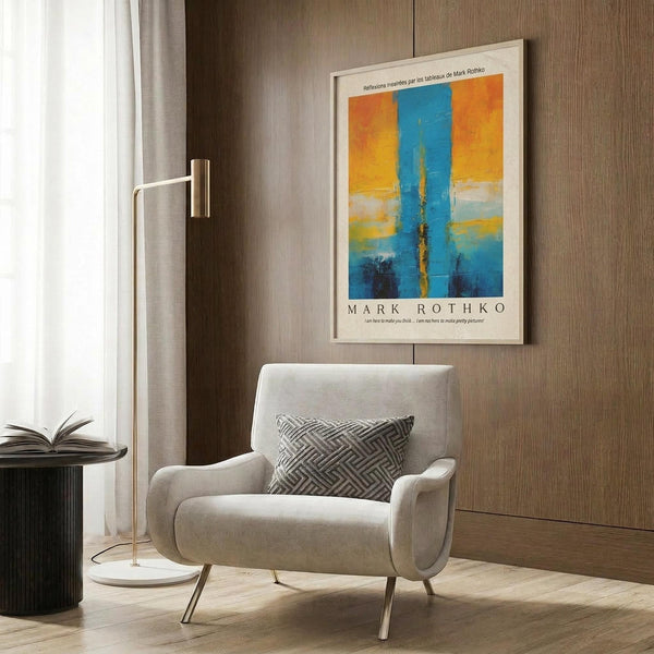 May include: Modern living room with beige armchair featuring geometric pillow, brass floor lamp, and framed abstract art poster on wood-paneled wall. The artwork displays vibrant blue and orange color blocks in Rothko's signature style with white matting and thin frame. Scene includes dark round side table with open book and white curtains.