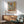 May include: Abstract art poster with geometric shapes in muted blues, oranges, and yellows displayed in thin wooden frame above mid-century modern credenza. Mint green upholstered armchair with wooden arms positioned nearby, white ceramic table lamp, warm natural lighting creating shadows on white wall.