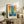 May include: Framed abstract art poster with blue vertical stripe through orange and yellow geometric blocks, displayed on rustic wooden sideboard next to fern plant, books, and ceramic vase in neutral living room setting with beige chair visible.