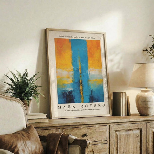 May include: Framed abstract art poster with blue vertical stripe through orange and yellow geometric blocks, displayed on rustic wooden sideboard next to fern plant, books, and ceramic vase in neutral living room setting with beige chair visible.