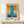 May include: Framed abstract art print with bold blue vertical brushstroke on warm orange and yellow background, displayed on light wood console table beside small white wrapped gift with twine and greenery, against cream wall in minimalist interior setting.