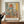 May include: Abstract art print in wooden frame displaying geometric shapes in muted orange, blue, and coral tones, positioned on wooden surface beside white ceramic vase and books. Person holds smartphone while viewing the framed artwork against cream-colored wall with white molding.