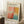 May include: Framed art poster with abstract colorful brushstrokes in coral pink, orange, yellow, and teal blue blocks. Features black text about an art gallery exhibition with dates April 15 - July 30, 2023. Light wood frame leaning against white wall corner on fluffy white rug. Small wooden block with white coffee cup and saucer nearby. Natural lighting creates soft shadows.
